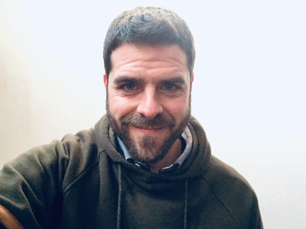 Hombre con barba y cabello corto oscuro sonríe frente a la cámara; lleva una sudadera verde con capucha y una camisa azul debajo, posando frente a una pared clara.