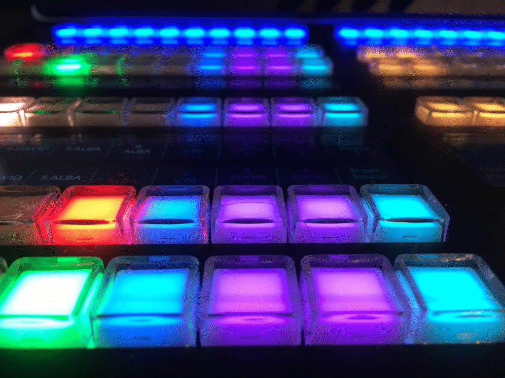 Primer plano de una consola de producción con botones retroiluminados en colores como verde, azul, morado, rojo y amarillo.