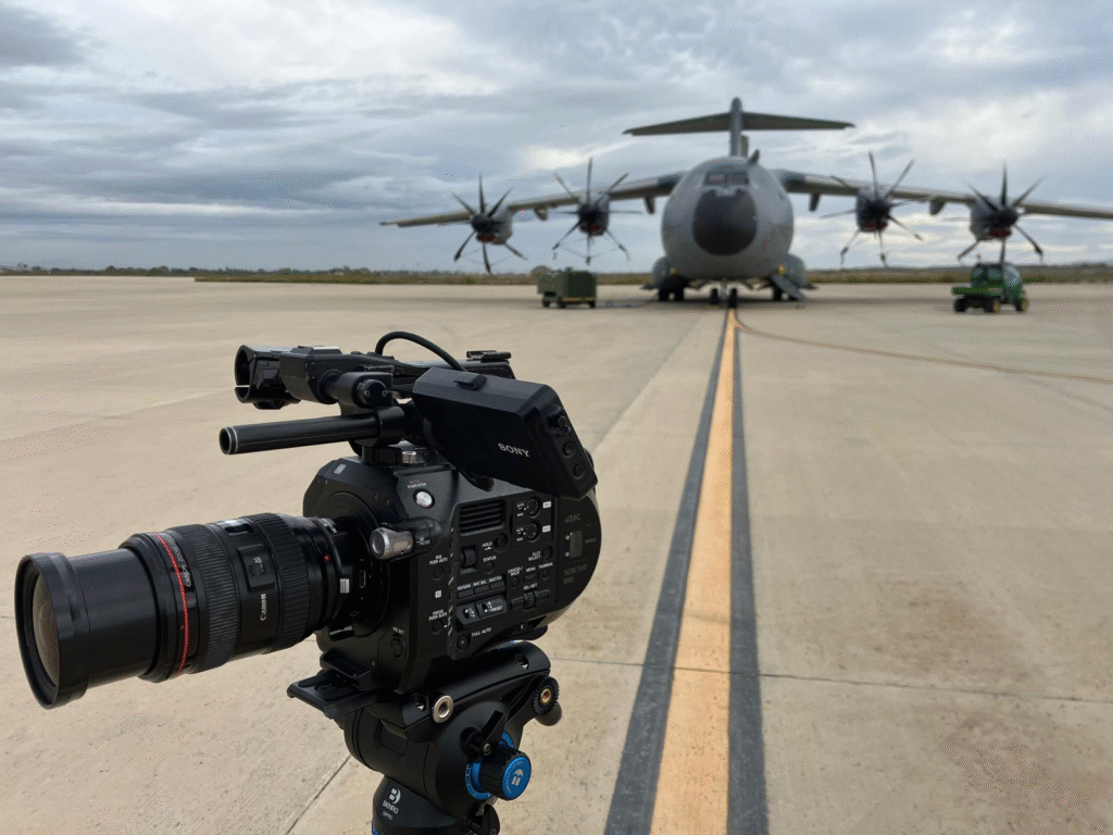 Cámara de video profesional montada sobre trípode enfocando un avión militar de gran tamaño con cuatro hélices, estacionado sobre una pista de concreto bajo un cielo nublado.