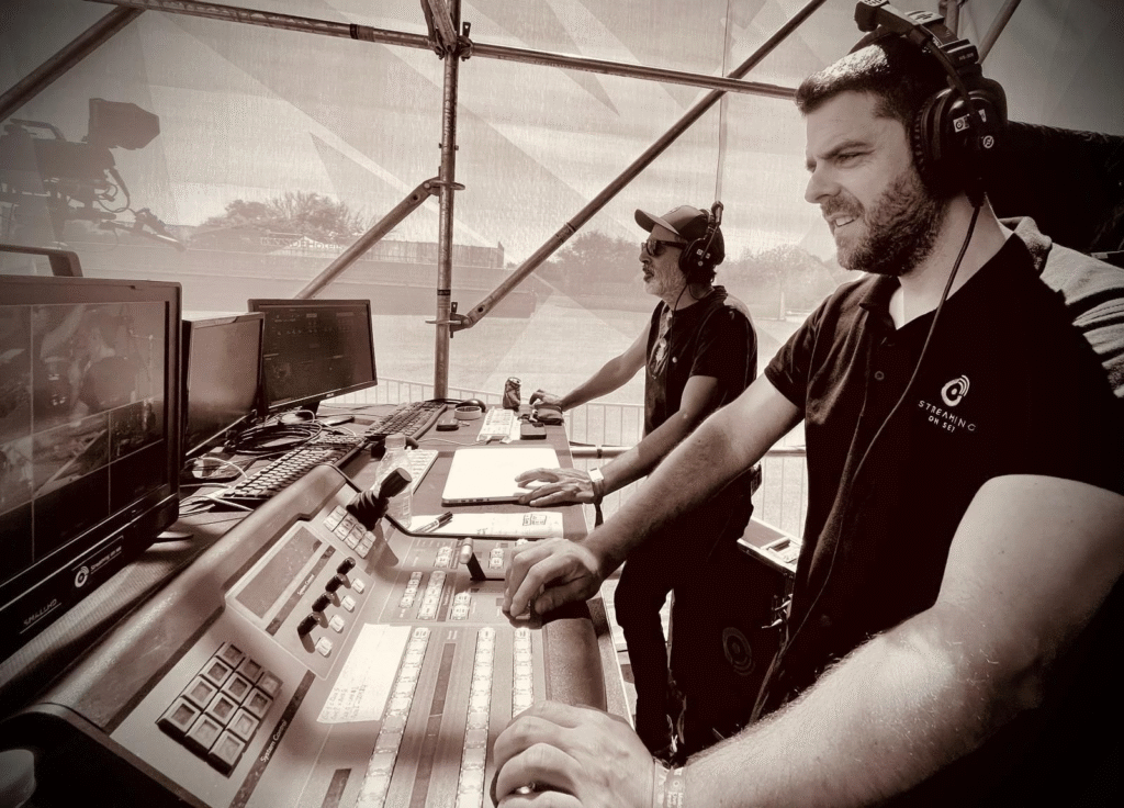 Dos hombres con audífonos operan una consola de transmisión con múltiples monitores en una cabina técnica al aire libre.